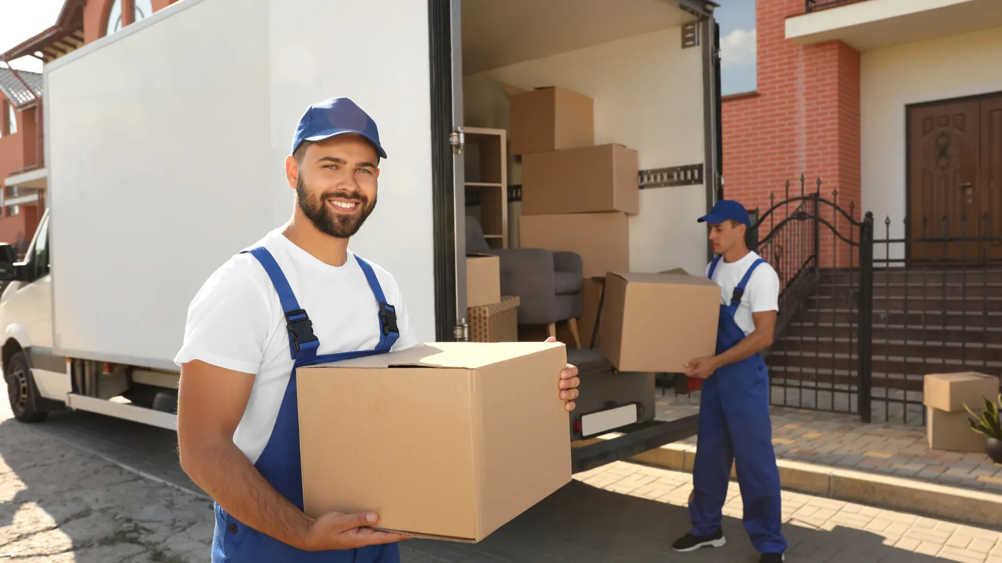 A smiling mover in a blue cap and blue coveralls over a white t-shirt looks at the camera while carrying a tan cardboard box